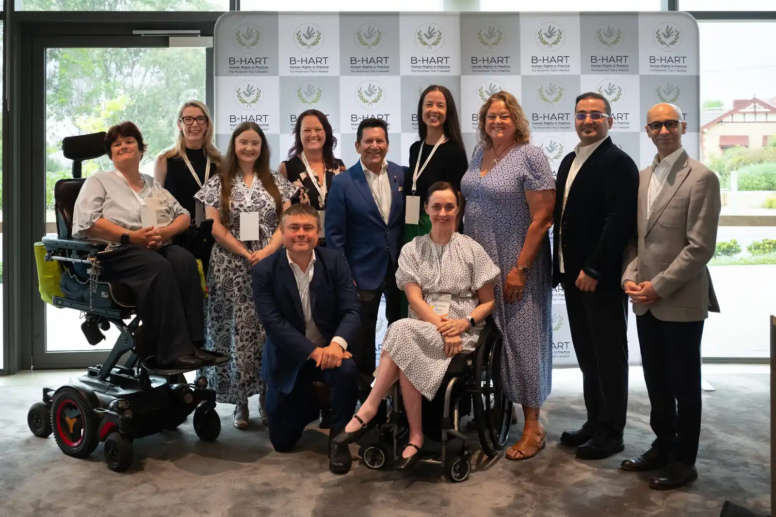 A large group of speakers and guests, including two people using wheelchairs, stand together in front of a B-HART banner.