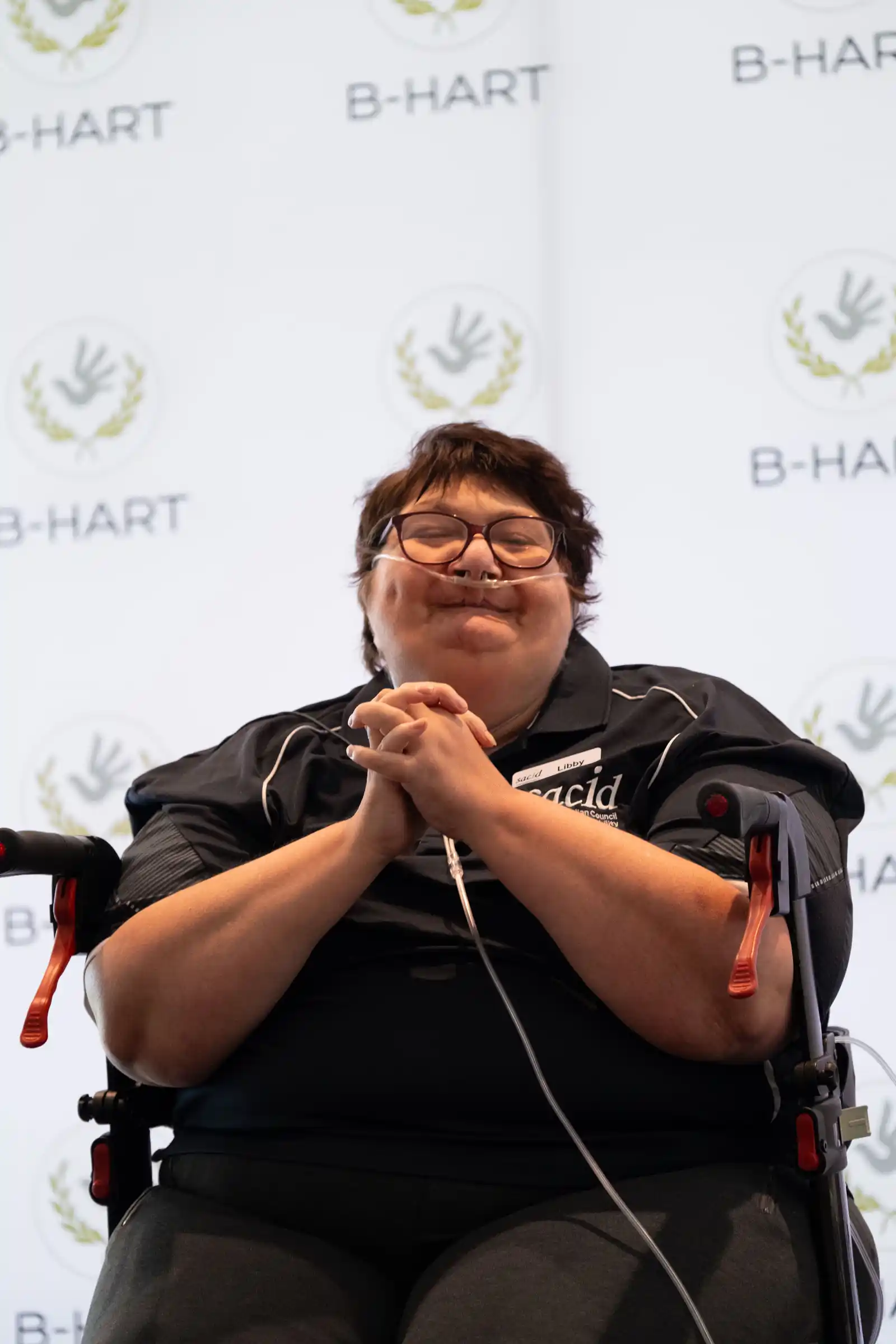A smiling woman using a wheelchair holds a microphone as she speaks in front of a B-HART banner.